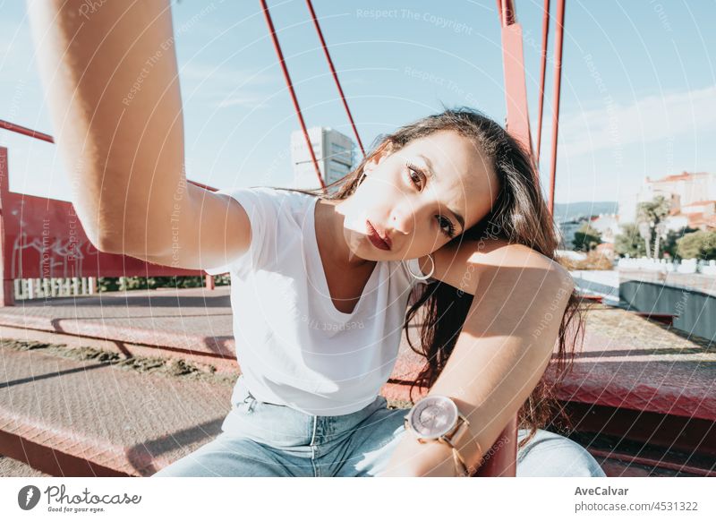 Young urban mixed race woman taking a selfie while looking serious to camera, urban lifestyle, white tshirt blank space, trendy social network, young people, wide angle image