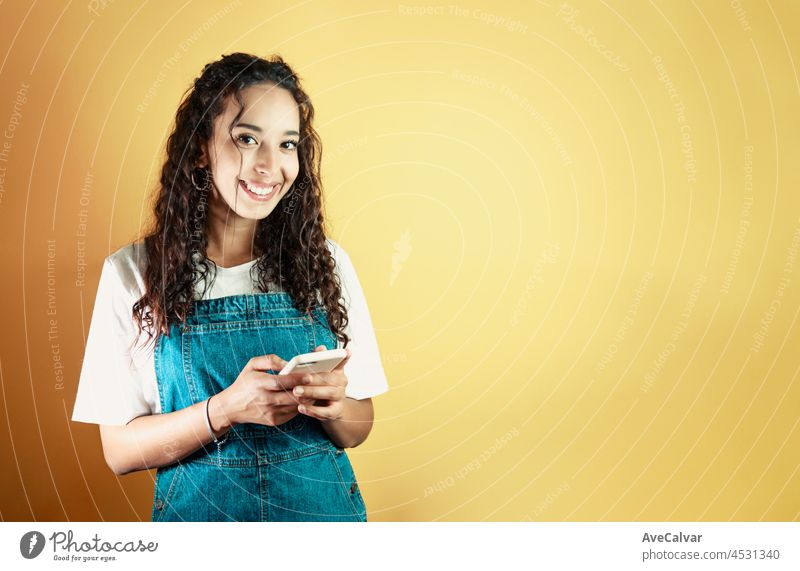 Young mixed race woman holding a phone texting a message and smiling to camera, yellow background removable background, minimal image, copy space, basic situations and feelings expressions
