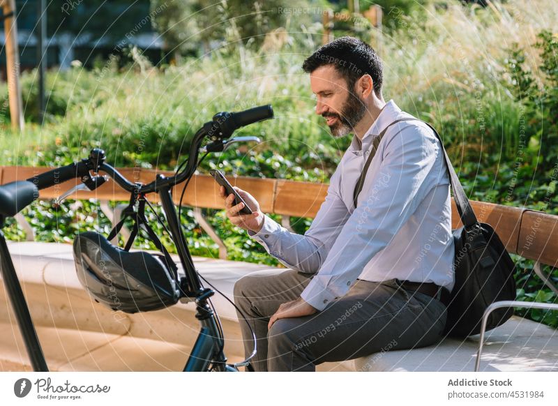 Serious man watching smartphone on bench in park manager using read check message bicycle male gadget browsing businessman employee text message cellphone