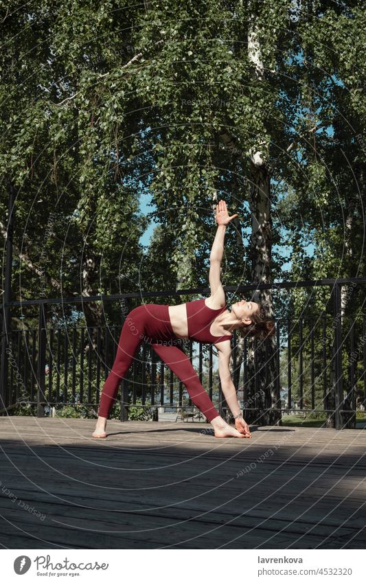 Young brunette woman practicing Trikonasana or Triangle pose in the park on wooden platform wellness body health lifestyle alone athletic balance casual