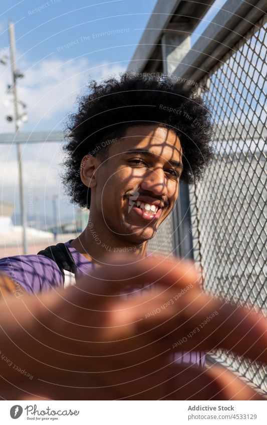 Smiling black man near net fence reaching hand to camera positive appearance carefree portrait pleasant shadow expressive glance grid male afro metal curly hair