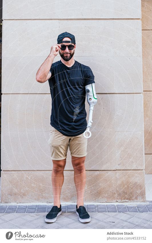Smiling hipster with arm prosthesis in city man artificial limb bionic metal cool male content handicap wall lean street smile young beard style urban building