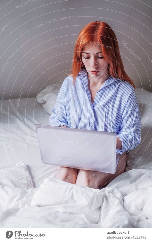 young woman sitting in bed using laptop on her knees doing remote work from home computer pajama notebook bedroom real people communication home office