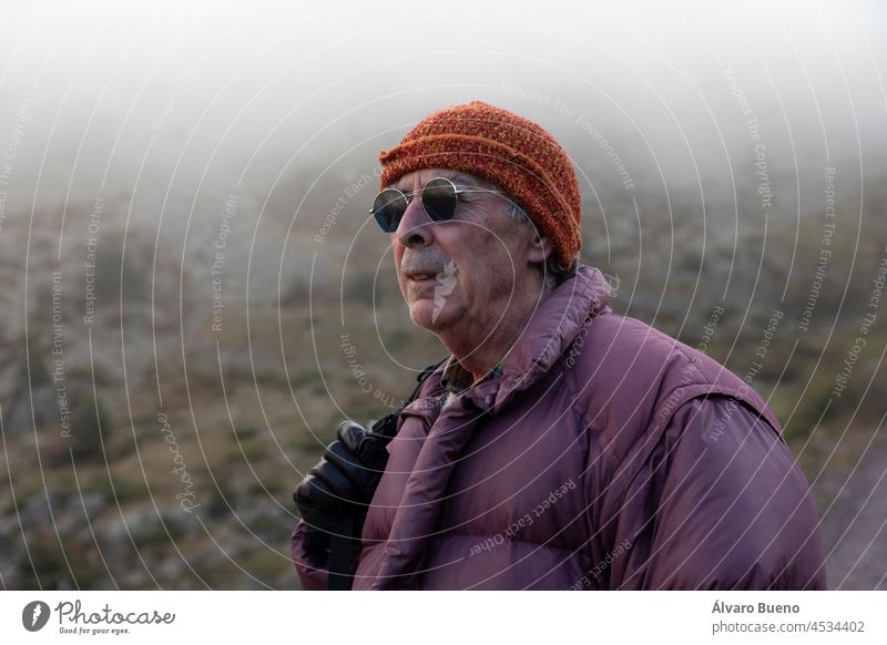 A mountaineer, senior adult man, in his 70s, wearing a down coat, gloves and sunglasses, enjoys the scenery during a mountain hike, Pyrenees, Spain 70 years old