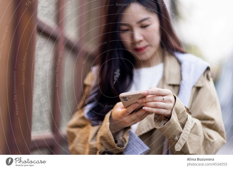 Stylish young asian woman dressed in casual wear looking to mobile phone screen when posting photos in social media and thinking about interesting content text. Lifestyle blogger checking blog comments outdoor at autumn