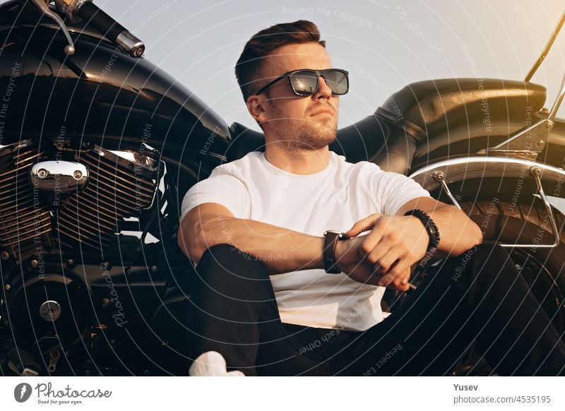 Young attractive guy of caucasian ethnicity sitting next to a motorcycle. Nice cute man in a in sunglasses, white T-shirt and black jeans. Life style photography.
