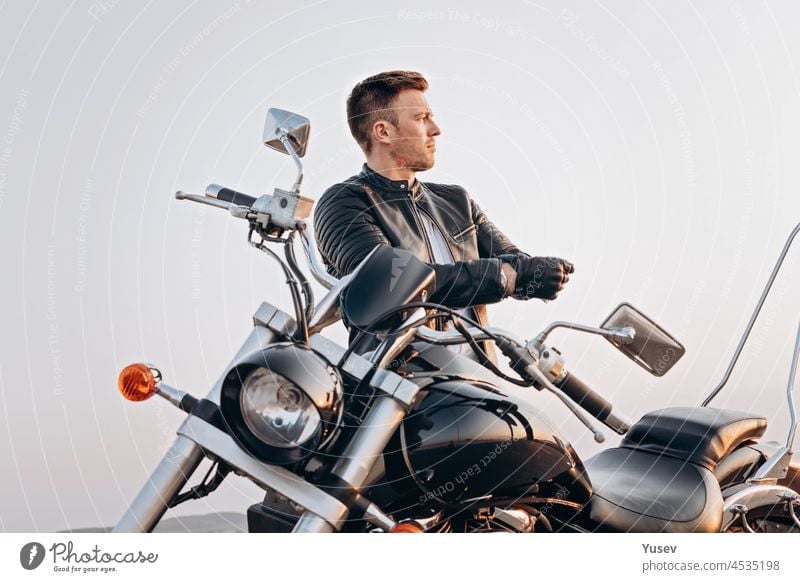 Attractive guy of Caucasian ethnicity in a leather jacket on a motorcycle. Portrait of a handsome young man on a black bike. Summer evening attractive stylish