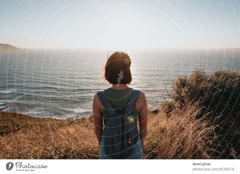 Back image of a young backpack traveler hiker during an stunning sunset coastline scenario, hipster traveler concept, beauty landscape. Copy space for text. Movement and liberty, nomad life.