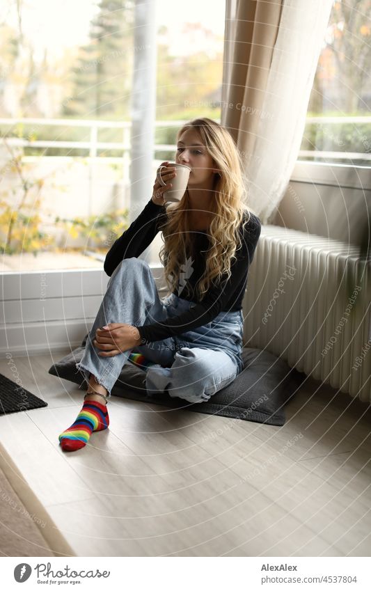 Young, slim, tall woman with long, blond hair sits in the living room in front of the balcony window and drinks from a big cup of tea or coffee Woman