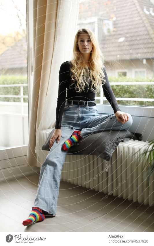 Young, slim, tall woman with long blond hair sits in the living room in front of the balcony window on the heater Woman Young woman Large Blonde Long-haired