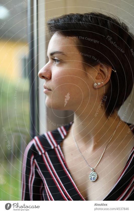 Young woman with short brunette hair looks out the window Woman short hair Brunette pretty naturally Authentic Jewellery Chain Earring Slim youthful 18-25 years