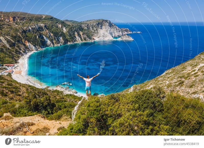 Man standing on top of a rock, raising hands with an exciting feeling of freedom, looking at Petani Beach. Cephalonia island, Greece beach myrtos aerial man