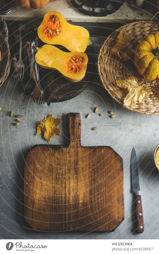 Food background with empty wooden cutting board on kitchen table with halved butternut squash, cutlery and kitchen utensils. Autumn cooking scene with seasonal ingredients. Top view.