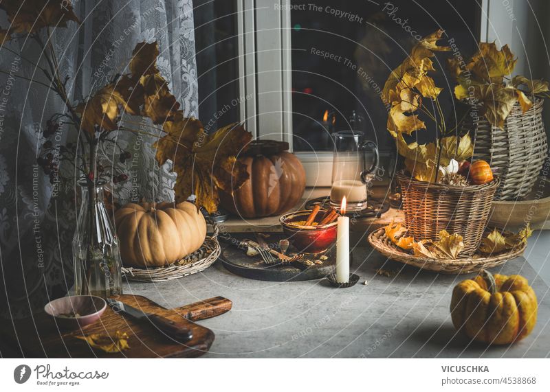 Cozy autumn kitchen background with pumpkins, autumn leaves, candles, cutting board and kitchen utensils on grey concrete table at window. Domestic still life. Front view.