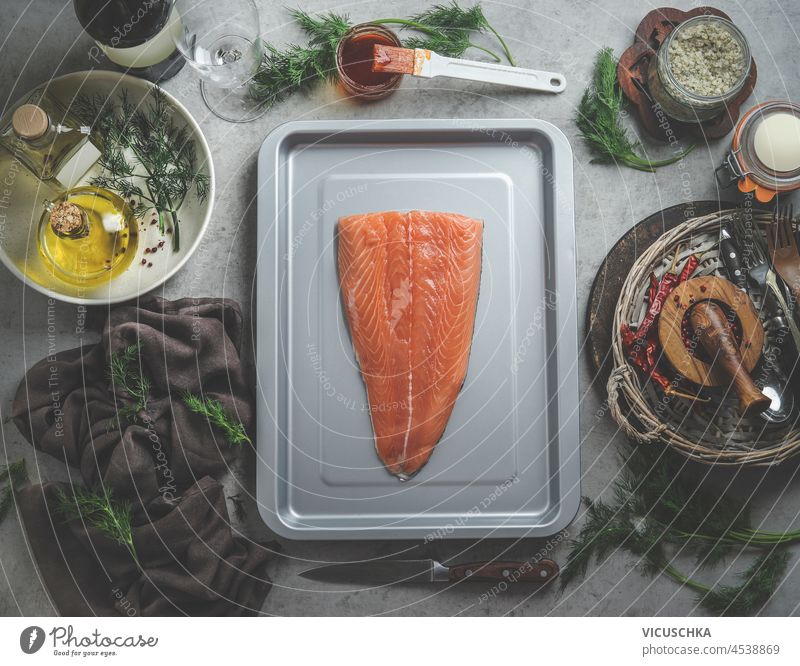 Raw salmon fillet on grey baking tray at kitchen table with herbs and spices, oil, brush with marinade, dish towel at grey kitchen table. Marinade fish at home with flavorful ingredients. Top view.