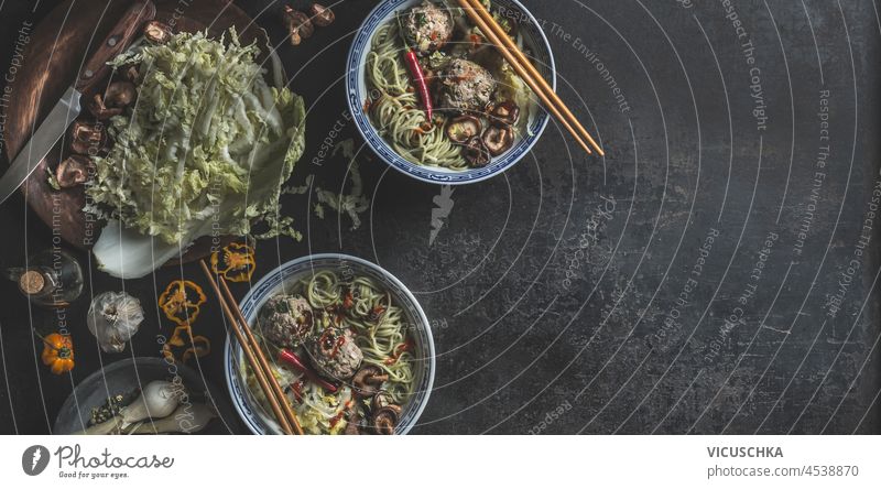 Bowls with Asian soup with noodle, cabbage, mushrooms, shiitake, garlic, chili and dumpling in traditional crockery with chopsticks on dark concrete table. Food banner with healthy ramen noodles. Top view