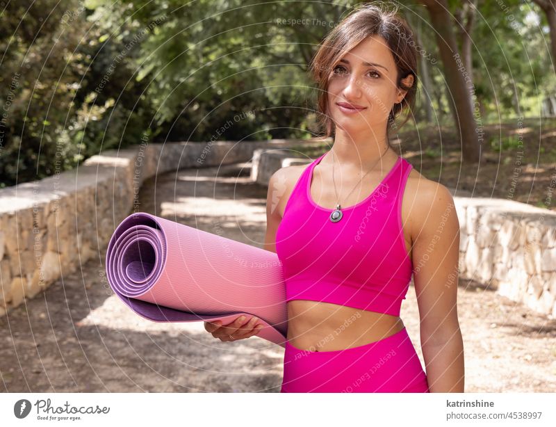 Smiling woman dressed sportswear with rolled pilates mat in a park stay mockup happy smile alone health summer pink green young outdoor fitness workout