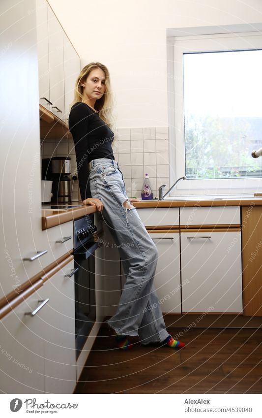Young, slim, tall woman with long, blond hair stands in a kitchen leaning against the cupboard and looks into the camera Woman Young woman Large Blonde