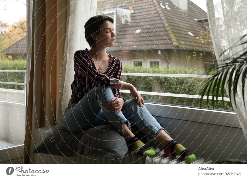 Young woman with short brunette hair sits on heater in front of window in living room Woman short hair Brunette pretty look naturally Authentic Jewellery Chain
