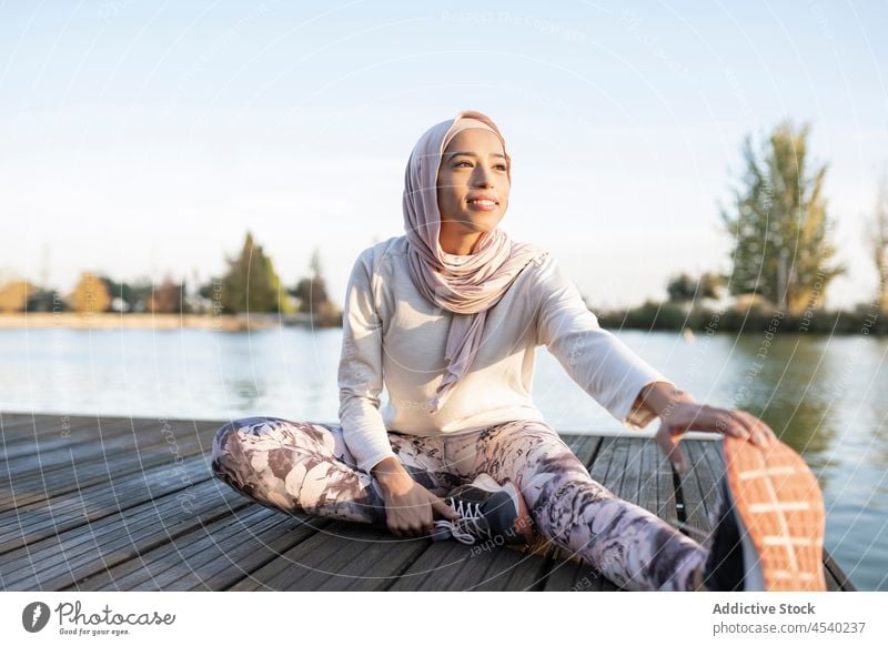 Positive Arab woman doing stretching exercise on embankment warm up waterfront river practice healthy lifestyle sport sporty workout female arab muslim