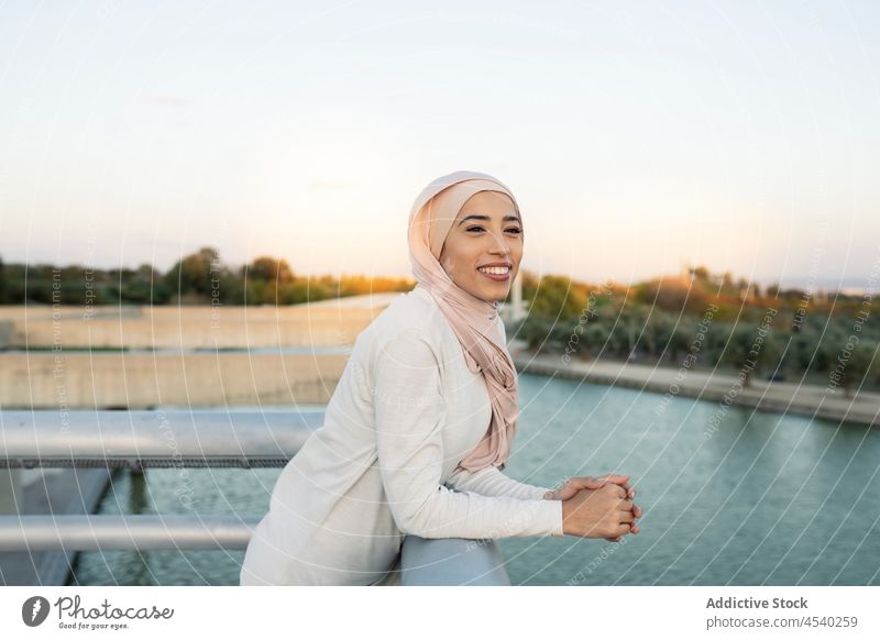 Positive Arab woman on embankment waterfront hijab river leisure pastime riverside shore railing female arab muslim lady summer fence smile content cheerful