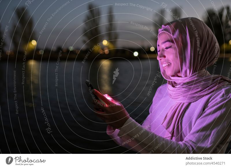 Positive Muslim woman using smartphone on embankment purple light sundown positive dark connection female tradition hijab sunset online mobile smile glow