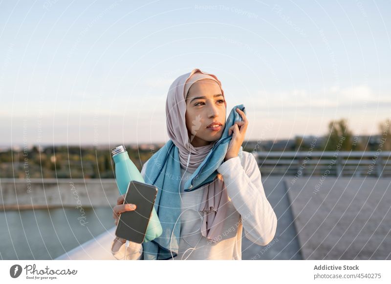 Tired Arab woman wiping face with towel after training tired sweat wipe listen music using earphones smartphone female workout muslim athlete bottle fitness