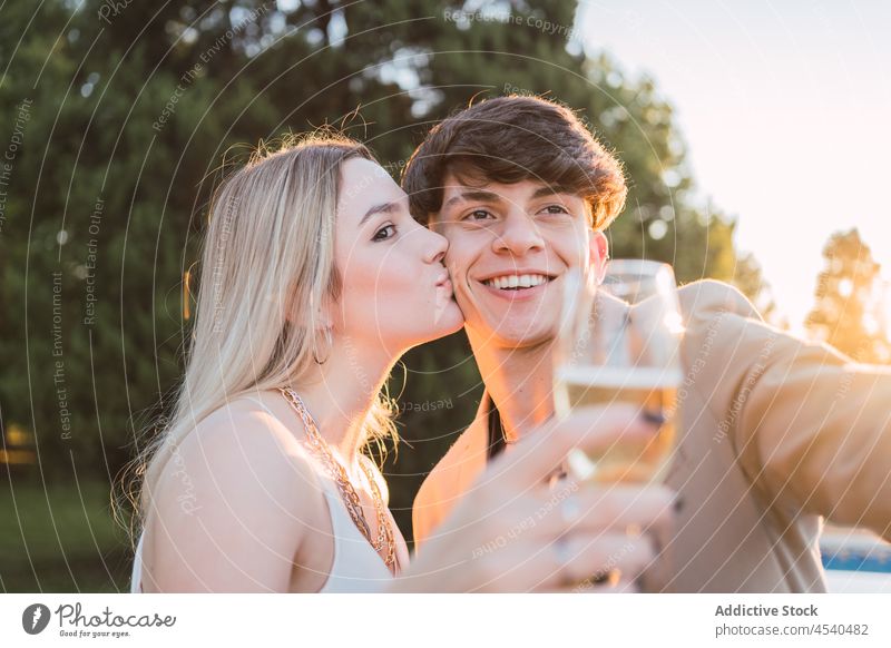 Woman with glass kissing partner for joint selfie in park couple cheerful alcohol together smartphone cellphone champagne self portrait carefree using chill