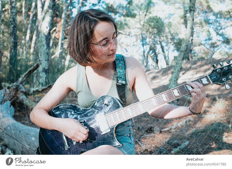 Young hipster woman playing the guitar outside the forest park of the city. Having fun learning a new skill, music play seasonal style. Young short hair girl. Copy space