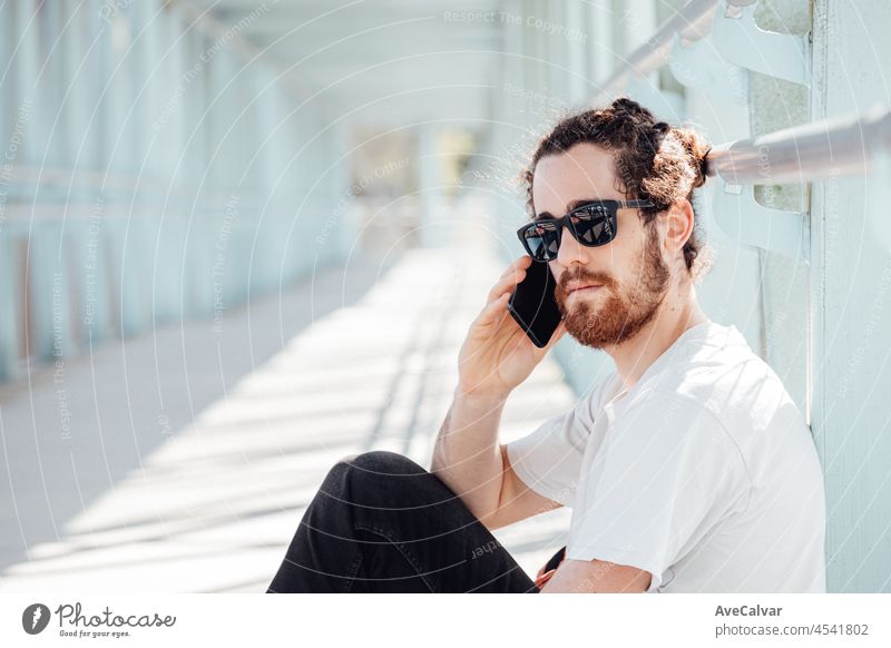 Young hipster man at the airport or bus station waiting while calls someone with the phone, luggage, bags and suitcase. Young man long hair hipster traveler with sunglasses, copy space, sunny day