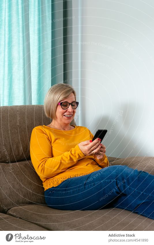 Smiling mature woman using smartphone on comfortable sofa internet digital connection positive chill pastime message female mobile gadget online eyeglasses