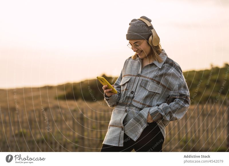 Happy young lady using smartphone and listening to music in headphones in countryside woman smile happy playlist meloman message hand in pocket nature female