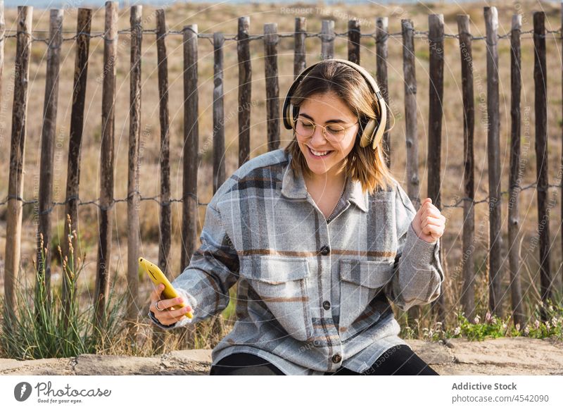 Happy young lady using smartphone and listening to music in headphones in countryside woman smile happy playlist meloman message nature female millennial trendy