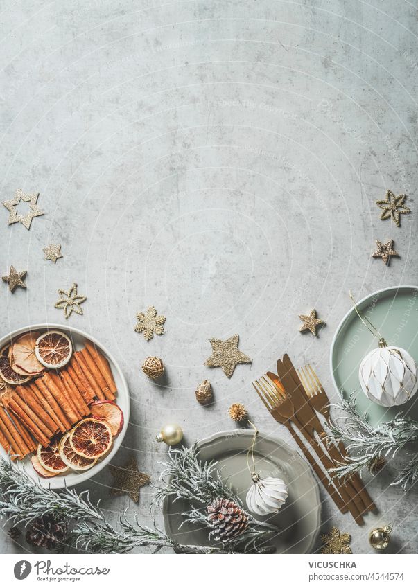 Christmas background with plates, gold cutlery, decoration, stars, cinnamon sticks, orange slices, bauble, pine cones and fir greens on pale grey concrete table. Top view with copy space.