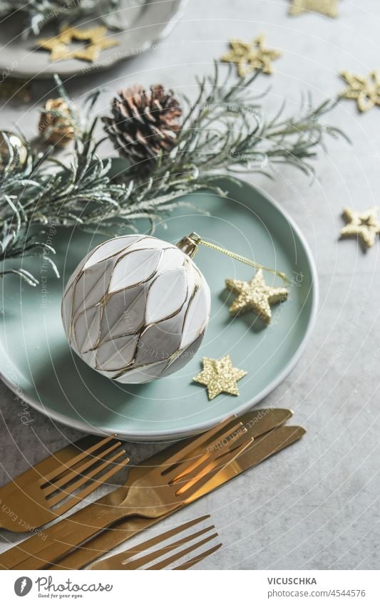 Christmas table setting with white vintage bauble, golden decoration stars, pine cone, golden cutlery, fir green and pale blue plate on grey concrete kitchen table. Top view.