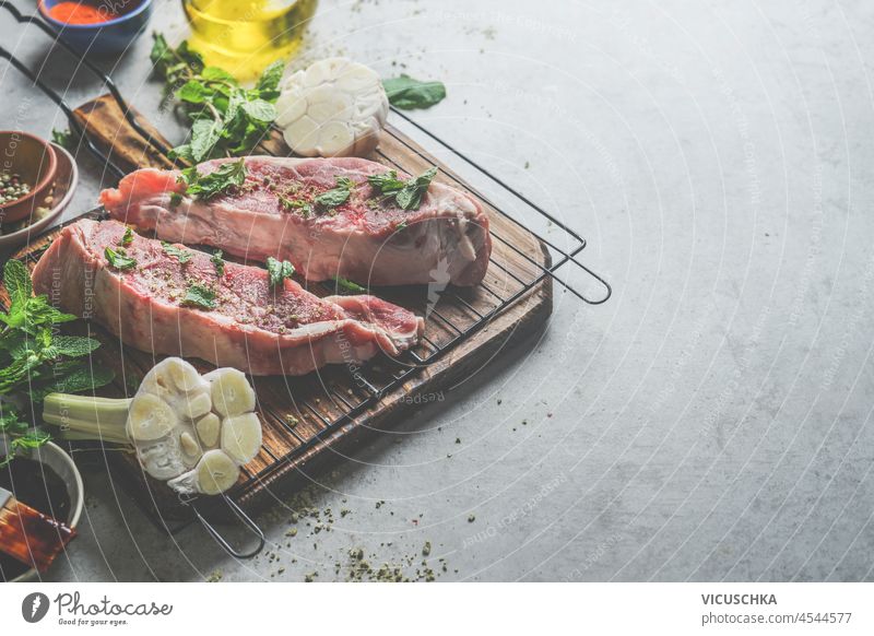 Raw meat on grill grate with flavor ingredients: garlic, herbs and spices on grey concrete kitchen table. Cooking preparation with steak for bbq. Top view with copy space.