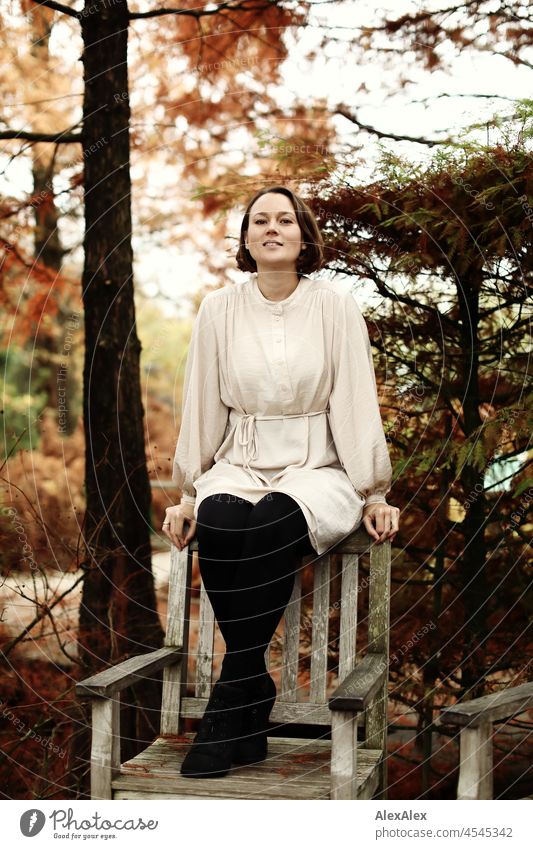 Young woman sitting on wooden chair back in park in autumn smiling at camera Cheerful Ease Beige Green Beautiful weather Moody Face of a woman free time