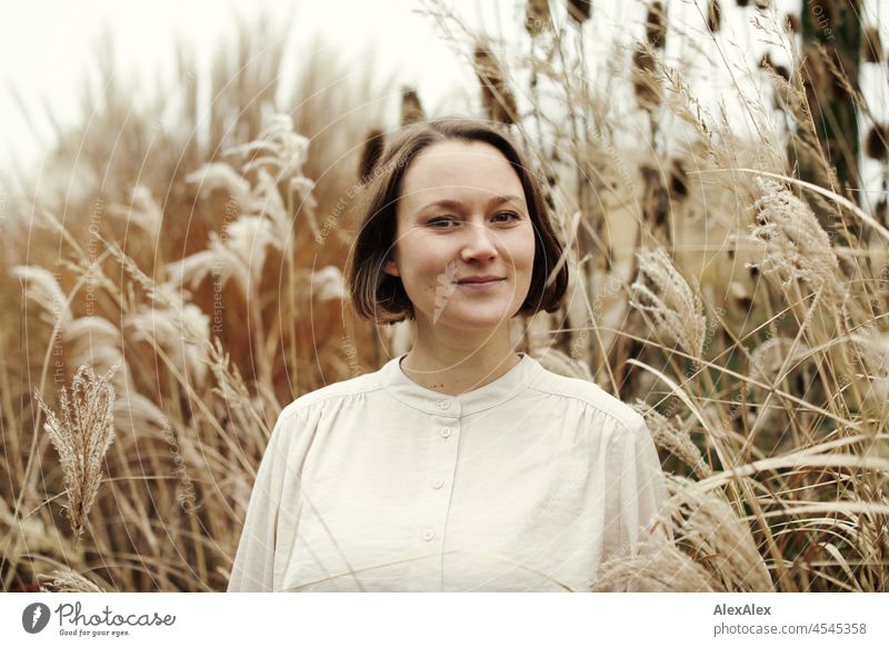 Portrait of a young smiling woman in the park in autumn in front of dry grasses Cheerful Ease Beige Green Beautiful weather Moody Face of a woman free time