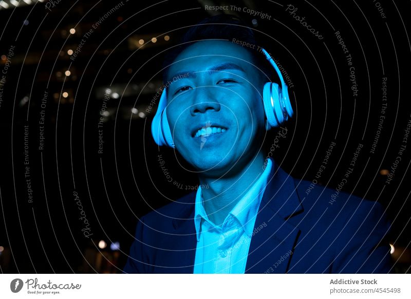 Cheerful Asian man in headphones on dark street music listen night song meloman formal pastime melody device audio asian darkness male positive cheerful enjoy