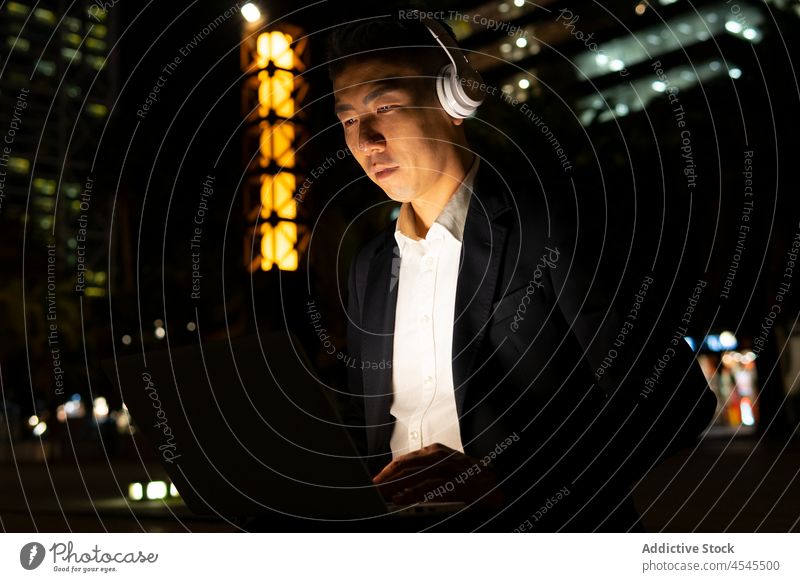 Asian businessman on laptop with headphones on dark street building night entrepreneur work cellphone remote suit outfit elegant respectable well dressed sound