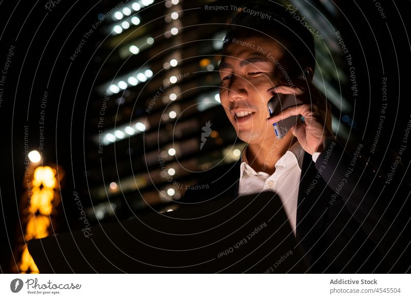 Asian businessman on laptop and talking on smartphone on dark street phone call conversation discuss building smile night happy communicate entrepreneur work