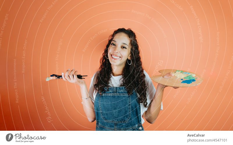 Young artist woman holding a palette and a paint brush over isolated orange background looking side. African curly hair woman. Art and creativeness concept, copy space. Smile