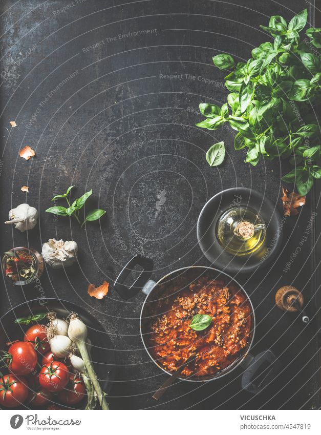 Sauce Bolognese in cooking pot at dark kitchen table with garlic, basil, tomatoes, olive oil and kitchen utensils. Cooking Italian recipe at home. Food background with copy space. Top view.