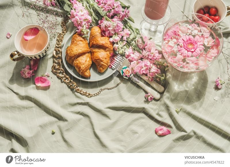 Summer picnic with french croissant, cherry blossom tea and fresh cherries on pale textile background. Romantic breakfast scene with pink petals, food and drinks. Top view.