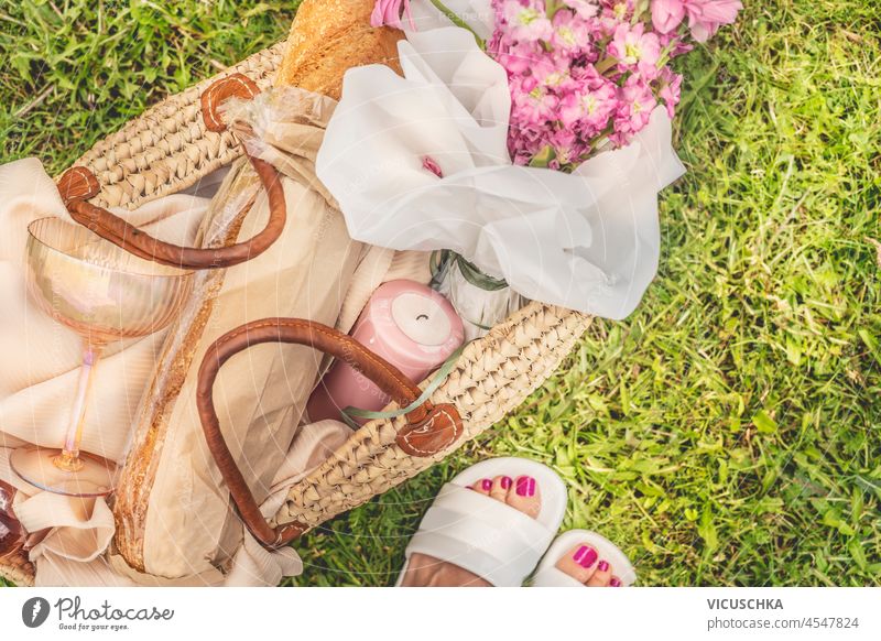 Picnic basket with candle, flowers, baguette and champagne glass on green grass background with woman feet. Romantic summer picnic preparation. Top view.