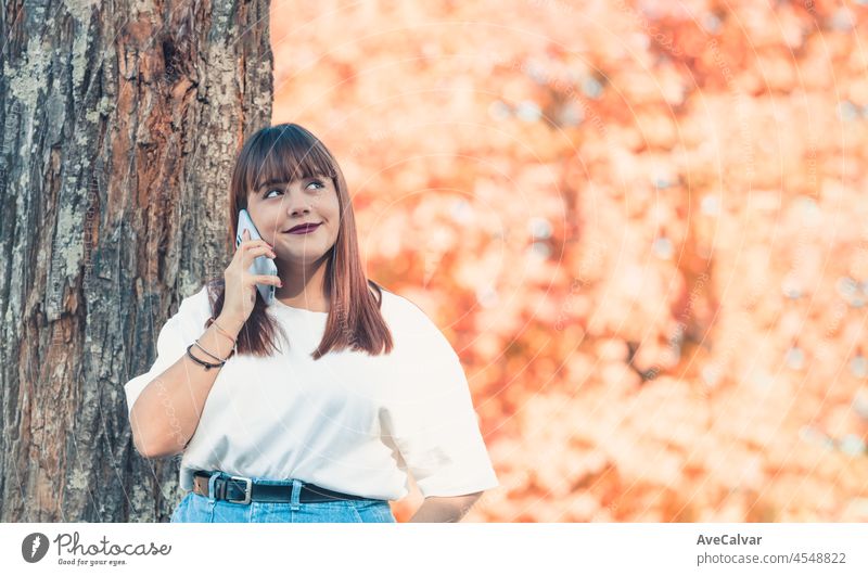 Young woman in park making a call using mobile phone on beautiful sunny day autumn. White tshirt clothes blank space. Redhead beauty body positive girl. Colorful background copy space