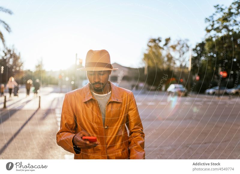 Stylish black man browsing smartphone on street online style fashion trendy text message connection african american hat surfing using serious outfit cellphone