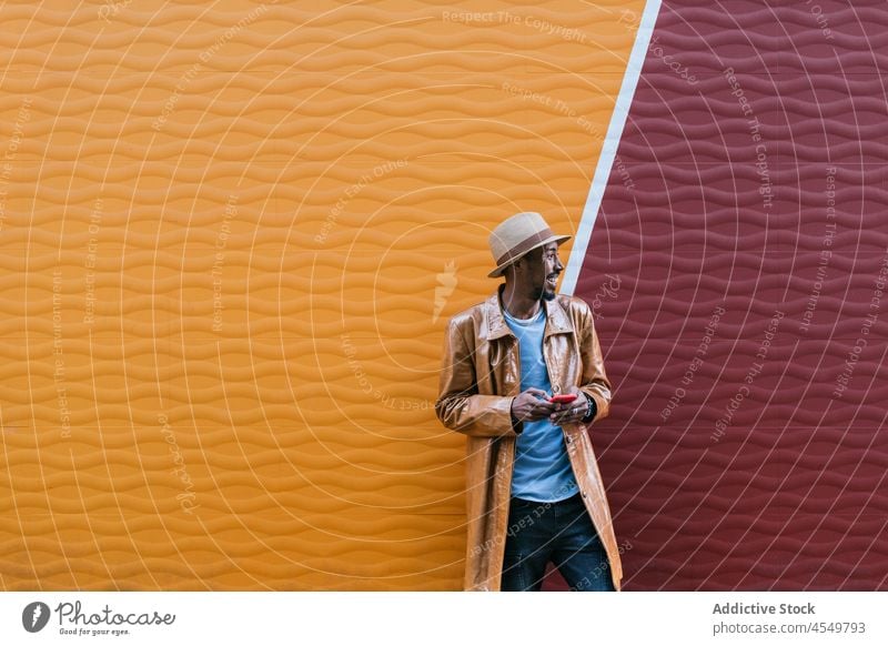 Cheerful black man browsing smartphone near wall online style street trendy text message connection african american hat surfing using outfit cellphone modern