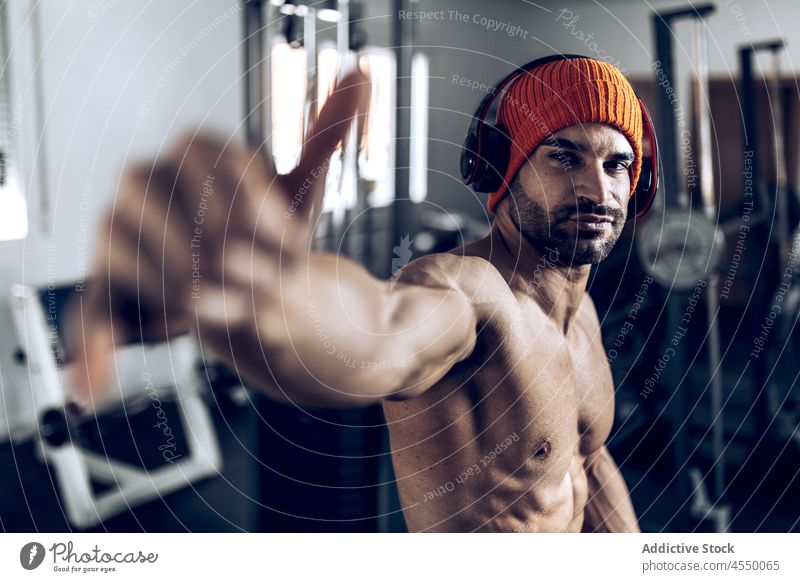 Ethnic sportsman showing hang loose gesture during training athlete shirtless shaka headphones strong muscular gym male listen workout hat fitness music