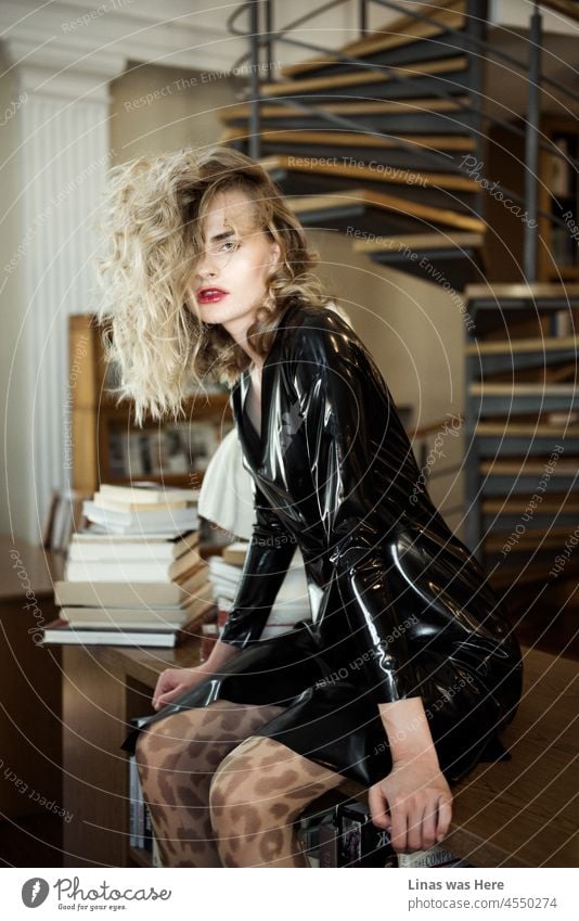 A gorgeous fashion model is posing with her black latex outfit. Her curly blond hair looks brilliant. Red lips are so sensual. The background seems to be a library.